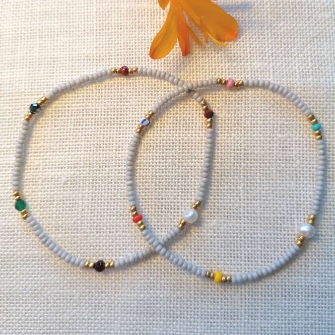 Elastikarmbånd, To lette armbånd med små lyse perler og diskrete farvepunkter, forbundet af et luftigt udtryk og fine gyldne afslutninger.