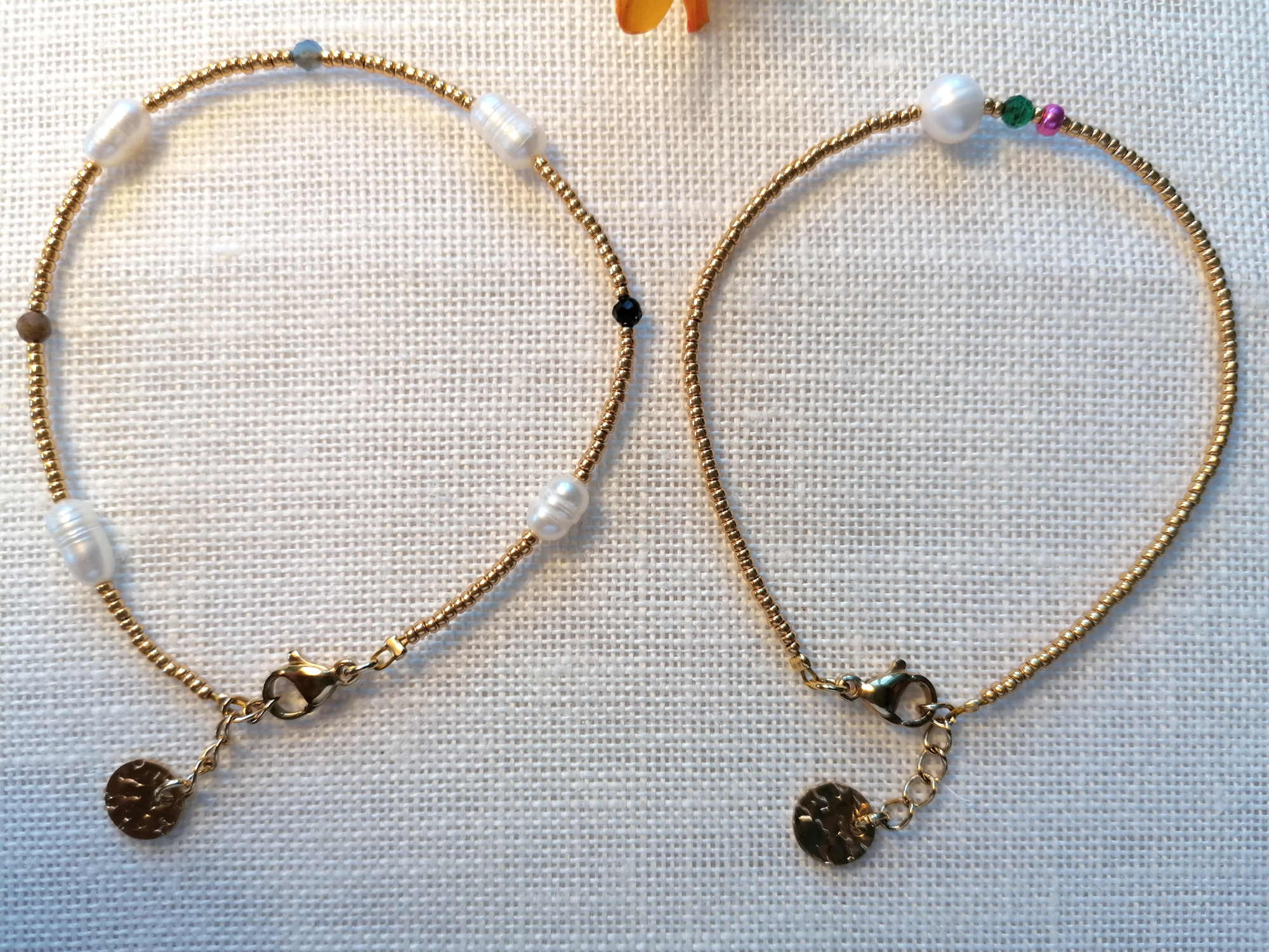 Armbånd, To fine armbånd i dæmpede naturtoner med enkelte farvede perler og små mørke vedhæng, hver med sit eget diskrete udtryk.